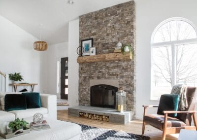 A white living room featuring a neutral stone fireplace with a natural wood banister. This room utilizes dark green throw pillows for a pop of color.