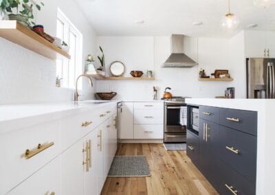 A white kitchen with gold hardware and open shelving. A Cutom built island features navy blue cabinets.