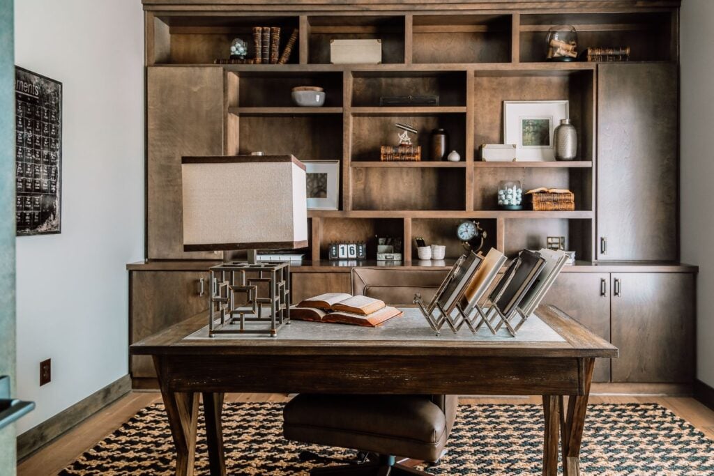 A rustic office with a wooden desk in the center. Behind the desk is a full wall of wooden shelves and cabinets. A brown and black patterned rug is used to create dimension in this room.