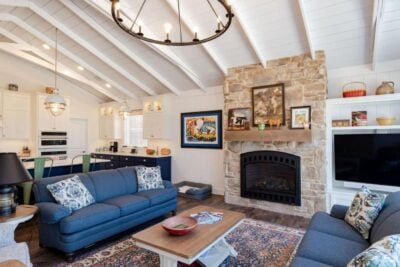 A cozy living room with a stone fireplace, wooden ceiling, and large chandelier. There's a blue sofa set, a wooden coffee table, and a TV. Shelves hold books and decor. The kitchen area is visible, featuring white cabinets and green barstools.