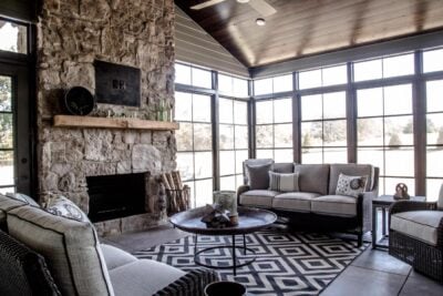 A cozy living room with large windows, a stone fireplace, and a wooden mantle. The space is furnished with gray sofas, a geometric patterned rug, and a round coffee table, creating a warm and inviting atmosphere.