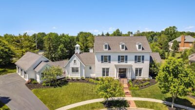Aerial view of a large, elegant white house with a gray roof, surrounded by a well-maintained lawn and trees. The home features multiple gables and a long driveway, under a clear blue sky.