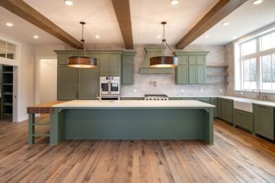 A spacious kitchen with green cabinets and a large central island. Wooden beams accent the ceiling, and pendant lights hang above. The room features hardwood flooring and large windows providing ample natural light.